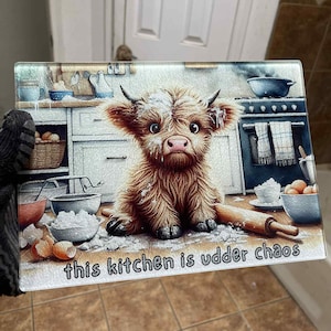 May include: Rectangular glass cutting board featuring a whimsical illustration of a Highland cow in a kitchen setting. The cow is surrounded by flour and baking supplies. Text on the board reads "this kitchen is udder chaos."