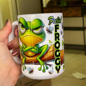 May include: A white ceramic mug featuring a cartoon green frog with a grumpy expression sitting on a brown log with its arms crossed. The text "Feelin' FROGGY" is printed vertically in green and black letters, surrounded by small flying insects.