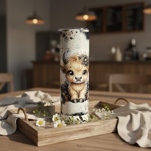 May include: A cylindrical stainless steel tumbler with a cartoon Highland cow design. The cow has large eyes and a flower crown, set against a white and black cowhide pattern. The tumbler has a silver lid and is suitable for hot or cold drinks.