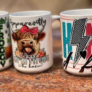 May include: Three white ceramic mugs featuring colourful graphic prints. The left mug shows a frog with the text Feelin' FROGGY. The centre mug displays a highland cow with a red bow and the text Apparently I have AN ATTITUDE Who knew?. The right mug has text.