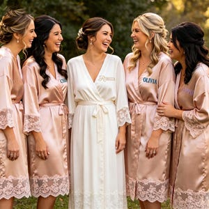 May include: A group of women wearing silk robes in shades of blush and ivory, with lace trim. One woman wears a white robe with the name "Rebecca" embroidered on it. The other robes have names embroidered on them as well.