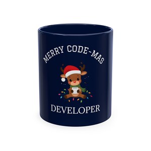 Peut inclure: Un mug en céramique bleu foncé orné d'un renne de dessin animé portant un bonnet de Père Noël rouge et une écharpe verte, tenant une tasse blanche. Le texte MERRY CODE-MAS est imprimé en blanc au-dessus et DEVELOPER est imprimé en dessous.