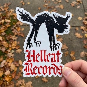 May include: A white sticker featuring a black silhouette of a cat with an arched back and a menacing expression. The sticker has the words "Hellcat Records" in red gothic lettering.