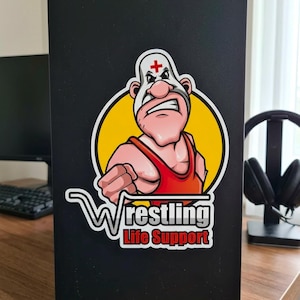 May include: A sticker featuring a cartoon wrestler with a red singlet and a white mask with a red cross. The wrestler is inside a yellow circle, with the words "Wrestling Life Support" below in white and red. The sticker is on a black surface.