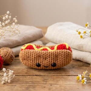 May include: A handmade crochet hot dog with a smiling face. The brown bun is filled with red and yellow yarn, resembling ketchup and mustard. The image includes yarn balls and flowers on a wooden surface.