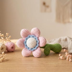 Könnte beinhalten: Eine handgefertigte Häkelblume in Rosa, Blau und Weiß mit einem grünen Stiel liegt auf einer Holzoberfläche. Garnknäuel und Trockenblumen deuten auf ein Bastel- oder Wohnkulturthema hin.