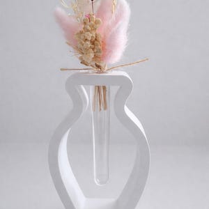 Könnte beinhalten: Eine moderne, weiße Vase mit einem einzigartigen, offenen Design, die einen kleinen Strauß Trockenblumen enthält. Der Strauß weist rosa, beige und weiße Elemente auf und erzeugt eine weiche, natürliche Ästhetik. Die Vase hat ein klares Glasröhrchen für die Blumen.