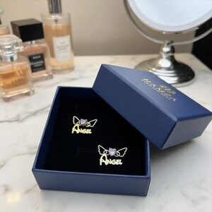 May include: A pair of gold-toned stud earrings in a navy blue box. Each earring features the word "ANGEL" in script lettering, topped with a pair of thin wire wings and a small, light purple heart-shaped stone centered between the wings.
