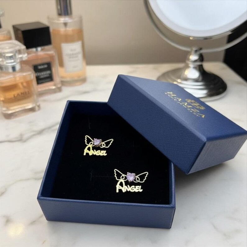 May include: A pair of gold-toned stud earrings in a navy blue box. Each earring features the word "ANGEL" in script lettering, topped with a pair of thin wire wings and a small, light purple heart-shaped stone centered between the wings.