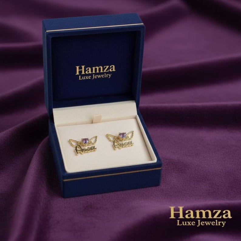 May include: A pair of gold-toned earrings featuring a heart-shaped purple stone and wing accents with the word Angel written in script, presented in a navy blue Hamza Luxe Jewelry box with a cream-colored interior and gold lettering on the lid.