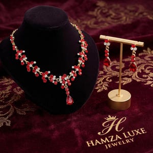 Puede incluir: Un conjunto de collar y pendientes colgantes en tono dorado con piedras rojas en forma de lágrima y pequeños cristales transparentes. El collar tiene un diseño de eslabones florales y los pendientes cuelgan de un soporte de metal dorado.