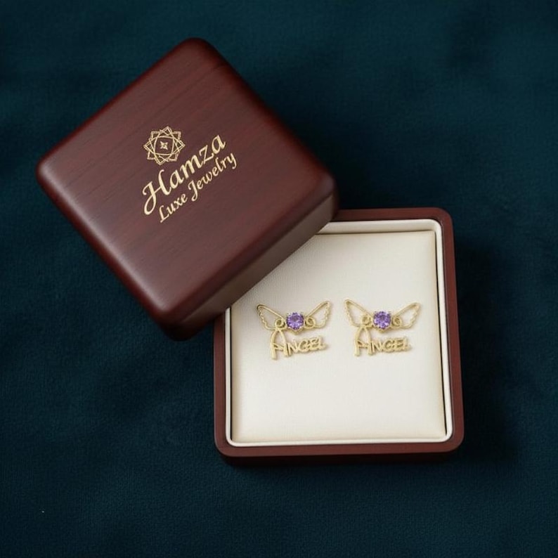 May include: A pair of gold-toned stud earrings featuring a purple circular gemstone, wing-shaped accents, and the word "ANGEL" in script lettering. The earrings are presented in a dark brown wooden jewelry box with "Hamza Luxe Jewelry" printed in gold on the lid.