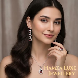 Op de afbeelding: Een sieradenset met een ketting, oorbellen en ring. De set bevat een hangerketting, oorhangers en een ring, allemaal met een druppelvormige, dieprode edelsteen omringd door kleine, heldere stenen. De tekst "HAMZA LUXE JEWELLERY" is zichtbaar.