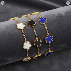 Op de afbeelding: Drie goudkleurige armbanden met klavervormige hangers in wit, zwart en blauw. Elke armband heeft een fijne ketting en een bijpassende klavervormige sluiting. De armbanden worden gepresenteerd op een grijze ondergrond.
