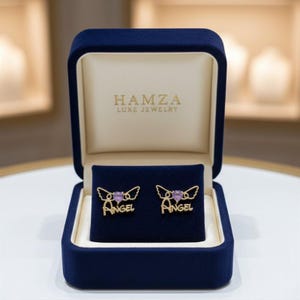 May include: A pair of gold-toned earrings featuring a purple heart-shaped stone, wings, and the word Angel in script, displayed in a navy blue velvet jewelry box with the text HAMZA LUXE JEWELRY printed in gold on the interior lid lining.