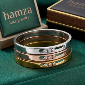 May include: A stack of three hinged bangle bracelets in silver, rose gold, and yellow gold tones. Each bracelet features a horizontal cutout slot containing three small, round, clear stones. The brand name hamza luxe jewelry is visible.