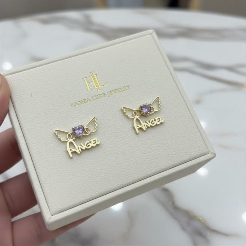 May include: A pair of gold-tone stud earrings featuring a small purple heart-shaped stone, stylized wings, and the word "ANGEL" in script lettering. The earrings are displayed on a cream-colored jewelry box branded with "HL HAMZA LUXE JEWELRY".