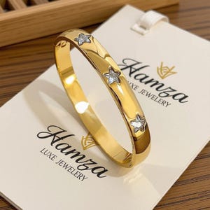 Może przedstawiać: Złota bransoletka typu bangle z trzema osadzonymi przezroczystymi kamieniami w kształcie gwiazdek. Bransoletka spoczywa na białej karcie ekspozycyjnej z nadrukiem Hamza LUXE JEWELRY. Metalowa powierzchnia jest polerowana, błyszcząca i zaokrąglona.