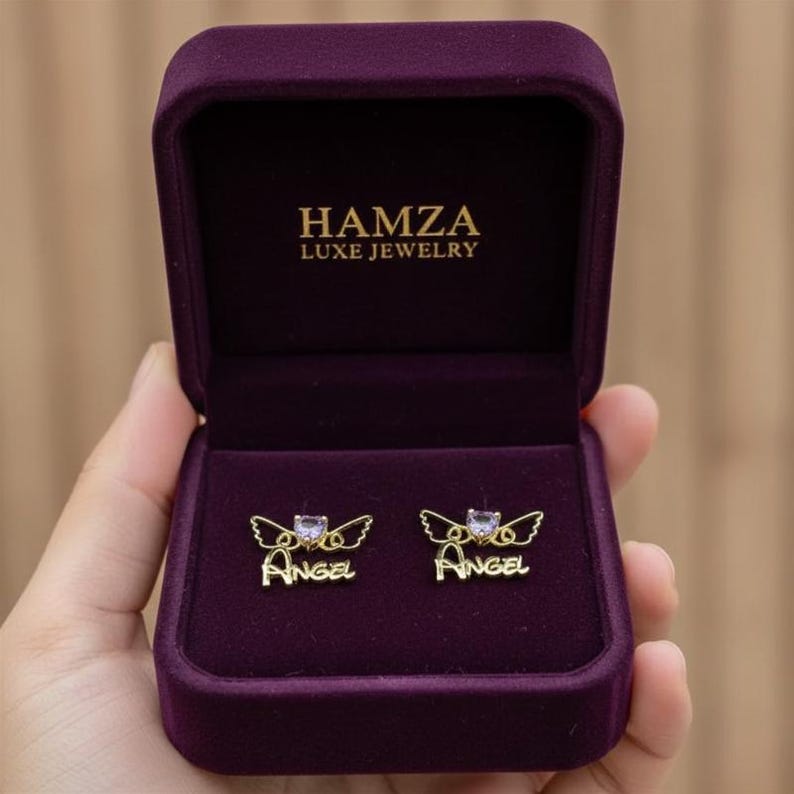 May include: A pair of gold-toned stud earrings featuring a heart-shaped stone, angel wings, and the word "Angel" in script, presented in a deep purple velvet jewelry box with the text "HAMZA LUXE JEWELRY" printed in gold on the inner lid.