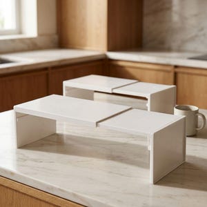 May include: Two white, rectangular, expandable shelf organizers with a smooth finish. Each unit features a flat top surface supported by two side panels, designed to create additional storage space by sliding to adjust the width as needed.