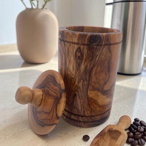 Könnte beinhalten: Ein zylindrischer Behälter aus Olivenholz mit passendem Deckel und kugelförmigem Griff. Das Holz weist eine markante, wirbelnde braune Maserung auf. Ein kleiner, passender Holzschöpfer liegt neben dem Behälter, begleitet von einigen dunklen Kaffeebohnen, die auf der Oberfläche verstreut sind.