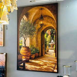 May include: A rectangular wall art print depicting a sunlit stone archway leading to a garden with a fountain. The scene features a hanging lantern, a potted olive tree in a large terracotta vessel, and warm golden light casting shadows.