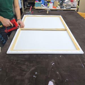 May include: A rectangular wooden canvas stretcher frame with a central support bar, featuring a white canvas stretched over the edges and secured with staples. A red pneumatic staple gun is positioned at the corner of the frame.