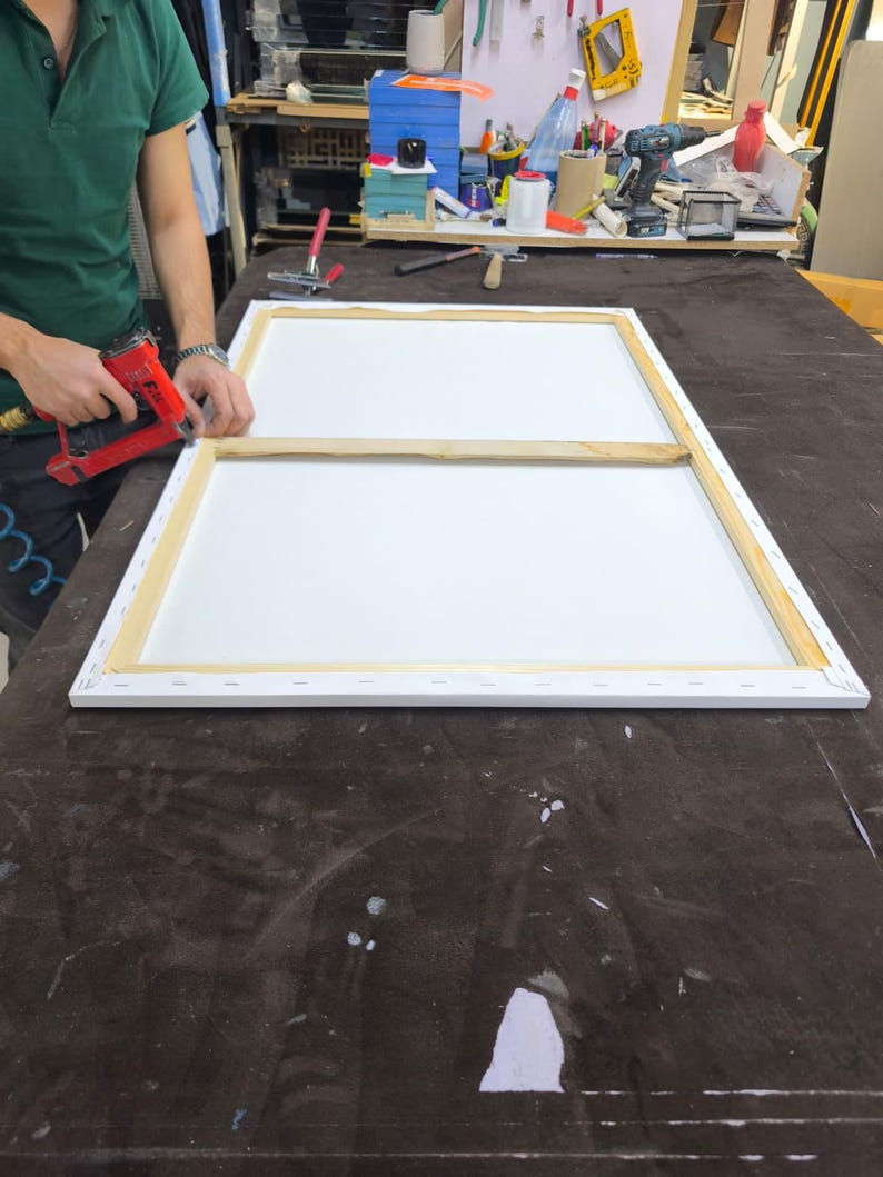 May include: A rectangular wooden canvas stretcher frame with a central support bar, featuring a white canvas stretched over the edges and secured with staples. A red pneumatic staple gun is positioned at the corner of the frame.