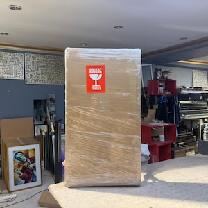 May include: A large rectangular cardboard box wrapped in clear plastic film. A bright red rectangular sticker is centered on the front, featuring a white wine glass icon and the text DIKKAT KIRILIR FRAGILE in white capital letters.