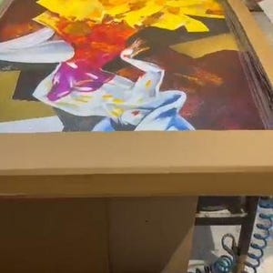 May include: A rectangular screen printing frame rests inside a cardboard box, featuring a vibrant graphic design with bold yellow, red, purple, and white abstract shapes. A blue coiled air hose is visible near the base of the frame.