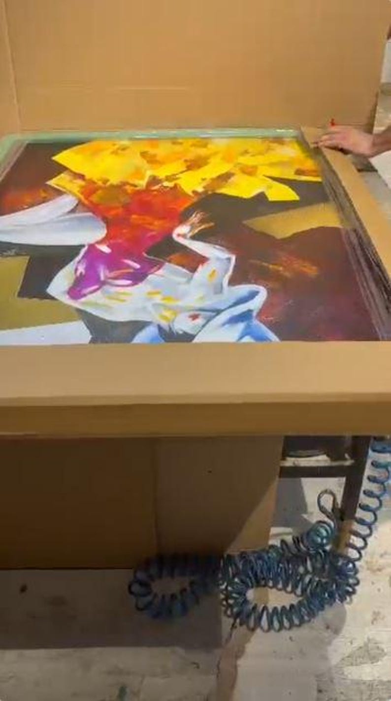 May include: A rectangular screen printing frame rests inside a cardboard box, featuring a vibrant graphic design with bold yellow, red, purple, and white abstract shapes. A blue coiled air hose is visible near the base of the frame.