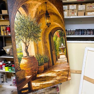 May include: A large rectangular canvas painting depicting a sunlit Mediterranean stone archway. The scene features a potted olive tree, a hanging black metal lantern, and a distant stone fountain. The color palette is dominated by warm golden, ochre, and earthy brown tones, with deep shadows and bright highlights creating a sense of depth and perspective.