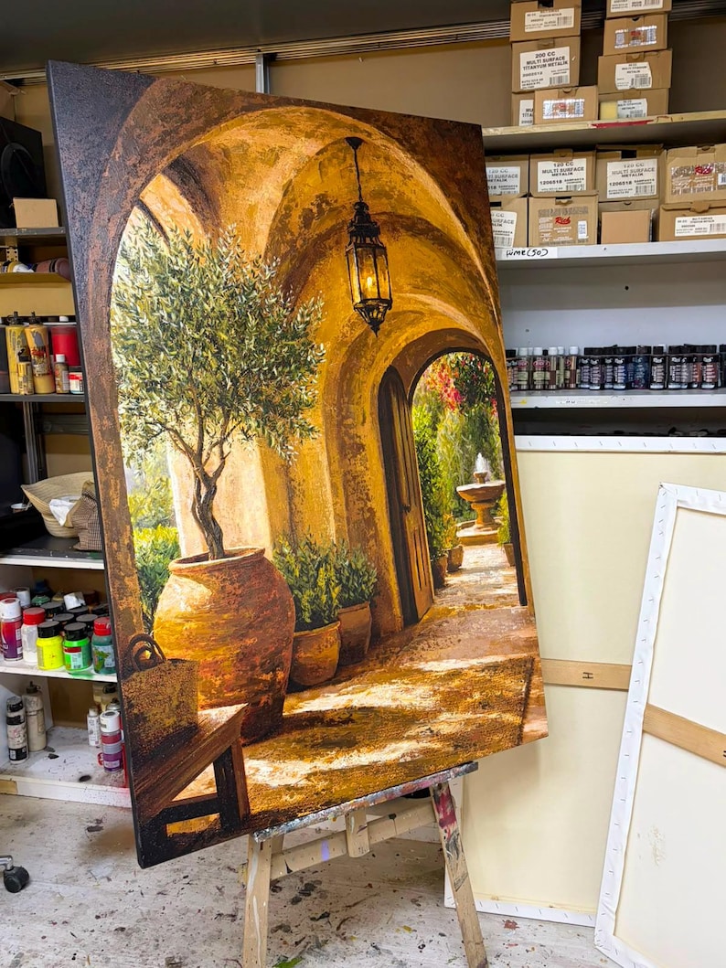 May include: A large rectangular canvas painting depicting a sunlit Mediterranean stone archway. The scene features a potted olive tree, a hanging black metal lantern, and a distant stone fountain. The color palette is dominated by warm golden, ochre, and earthy brown tones, with deep shadows and bright highlights creating a sense of depth and perspective.