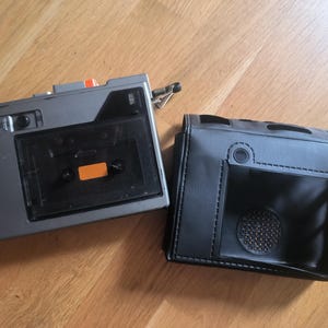May include: A gray portable cassette player with a clear plastic window and an orange label on the cassette tape, accompanied by a black faux leather protective carrying case featuring a circular speaker grille cutout and stitched detailing.