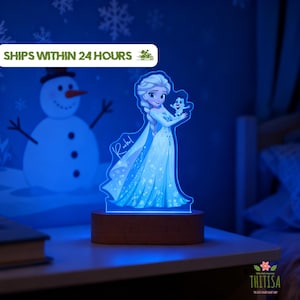 Könnte beinhalten: Ein LED-Nachtlicht aus Acryl mit einer blau getönten Illustration einer Figur mit langem blonden Zopf in einem Kleid, die eine kleine Schneemannfigur hält. Der Name Rachel ist an der Seite eingraviert. Das Licht steht auf einem Holzsockel.