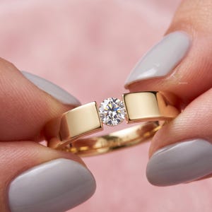 Puede incluir: Un anillo de oro con un diamante blanco único de talla brillante redonda, engastado entre dos bandas de oro lisas y pulidas. El metal tiene un acabado de alto brillo y la piedra central está asegurada en un engaste de tensión, creando un diseño limpio.
