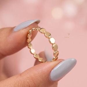 Könnte beinhalten: Ein goldener Ring mit einer Reihe kleiner, gleichmäßig verteilter, kreisförmiger Akzente. Der Ring wird vor einem weichen, rosa Hintergrund gehalten. Das Design des Rings ist schlicht und elegant.