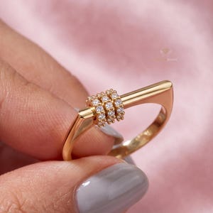 May include: A gold-toned ring featuring a horizontal bar design across the top. The center of the bar is adorned with three stacked, cylindrical bands encrusted with small, clear, sparkling stones that catch the light, creating a textured look.
