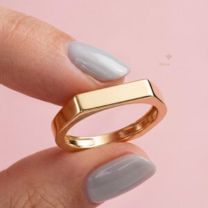 May include: A gold-colored ring with a rectangular top and a smooth, polished finish. The ring is held between two fingers against a soft pink background. The ring's design is modern and minimalist.