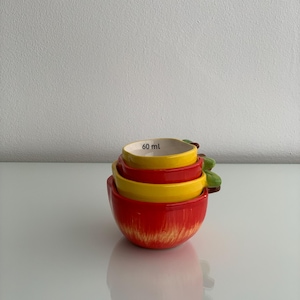 May include: A set of three nested ceramic measuring cups shaped like apples. The cups are coloured in shades of red and yellow with green leaf accents. The interior of the top cup is white and clearly displays the measurement text 60 ml in black.