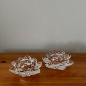 May include: Two clear glass lotus flower candle holders. The candle holders are made of clear glass and have a detailed lotus flower design. They are sitting on a wooden surface.