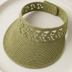 May include: An olive green crochet visor featuring a dense, circular-stitched brim and an openwork headband detailed with a repeating leaf and berry pattern. The back of the headband includes a hook-and-loop fastener for adjustable sizing.