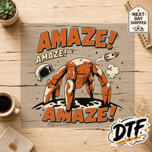 Peut inclure: Une impression graphique représentant un robot orange stylisé à quatre pattes debout sur une surface lunaire, accompagné d'un casque d'astronaute, d'une petite fusée et du texte répété AMAZE! en lettres orange gras avec des contours noirs.