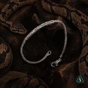 Könnte beinhalten: Ein silbernes Armband mit einem verzierten, gravierten Design. Das Armband hat einen detaillierten Verschluss und eine geflochtene Kette. Der Hintergrund zeigt eine Schlange mit braunen und schwarzen Mustern.