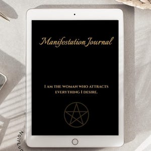 Può includere: Un tablet bianco mostra uno schermo nero con testo dorato: "Manifestation Journal". Sotto, c'è scritto "I AM THE WOMAN WHO ATTRACTS EVERYTHING I DESIRE" e un pentagramma dorato. Il tablet è su una superficie grigio chiaro.