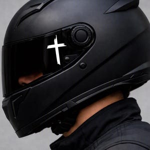 May include: A matte black full-face motorcycle helmet featuring a dark tinted visor. A white graphic of a cross with a distressed, brush-stroke texture is applied to the side of the visor, positioned near the pivot mechanism of the helmet.