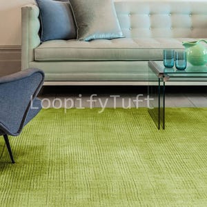 May include: A lime green textured rug with a ribbed design dominates the foreground. A light green sofa with two pillows, a glass coffee table with two blue glasses, and a blue chair are in the background. The text "LoopifyTuft" is visible.