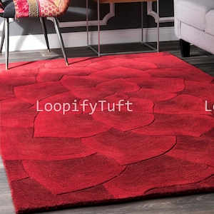 May include: A vibrant red area rug with a floral petal design. The rug features a textured surface and the text "LoopifyTuft" in white. The rug is placed on a wooden floor, with a chair and table in the background.