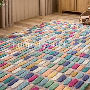 Peut inclure: Un tapis rectangulaire avec un motif texturé multicolore. Le tapis présente des rangées de formes oblongues dans diverses couleurs, notamment rose, vert, bleu, jaune et orange. Il est posé sur un sol en bois, créant une ambiance lumineuse et accueillante.