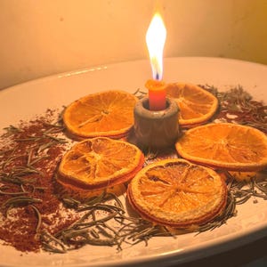 Puede incluir: Una vela naranja encendida en un portavelas de cerámica gris se encuentra sobre un plato blanco rodeado de rodajas de naranja secas, ramitas de romero y polvo marrón. La llama de la vela brilla intensamente.